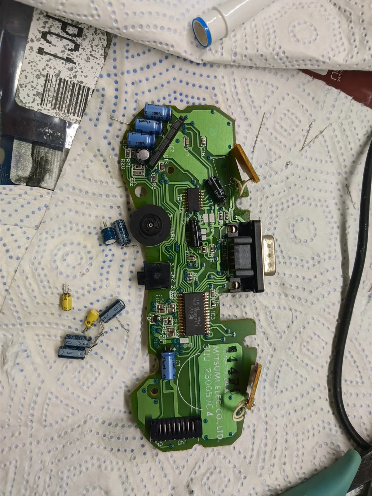 Disassembled 3DO controller PCB with removed capacitors laid out separately, blue tape, solder iron, and documentation during recapping repair.