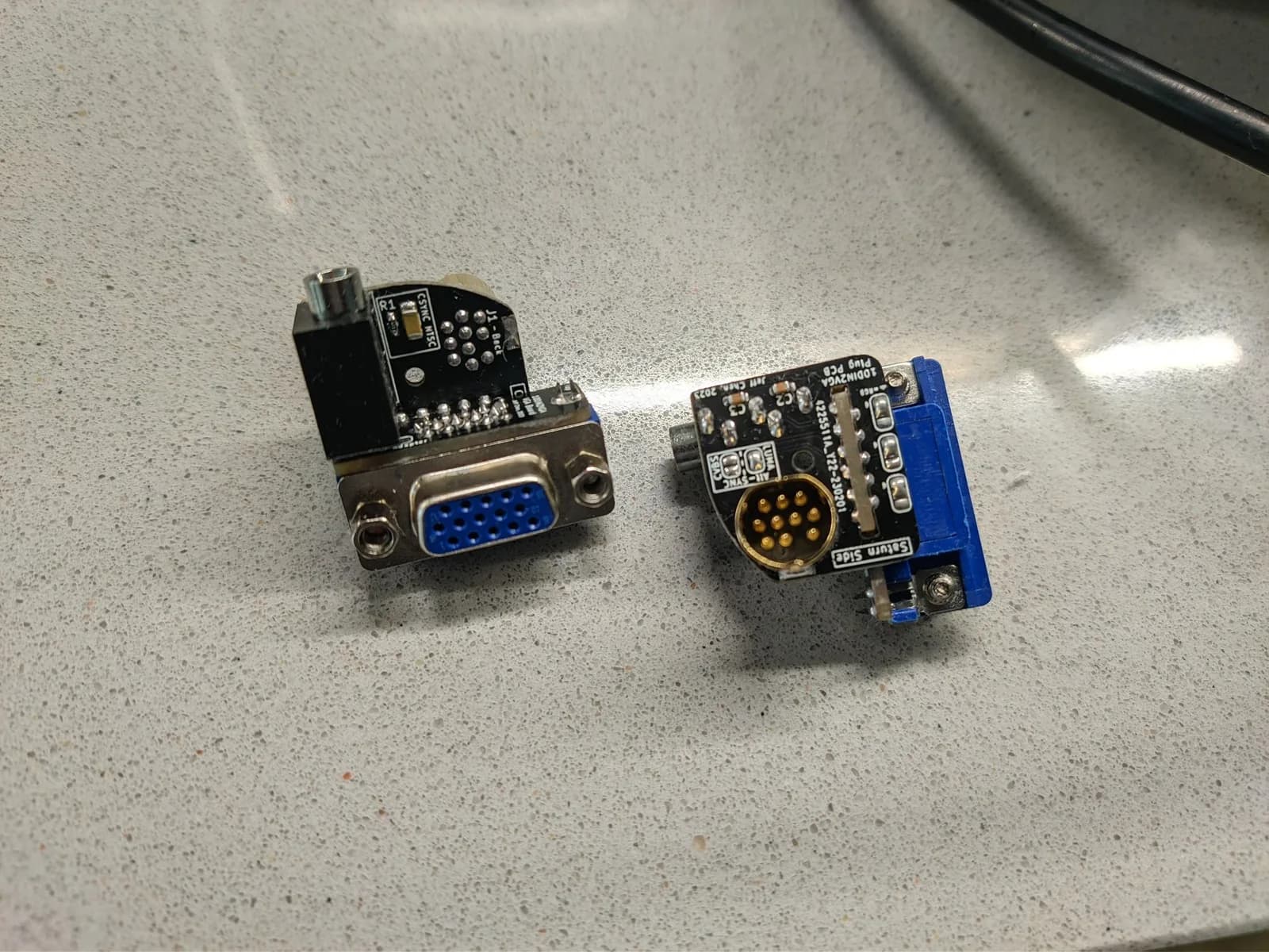 Two 10DIN2VGA (Saturn) dongle boards on a speckled stone worktop: the left one (configured for NTSC consoles) shows its blue 15-pin VGA connector on a black board; the right one (configured for PAL consoles) shows the gold 10-pin DIN Saturn-side connector (silkscreen '10DIN2VGA Plug PCB / Saturn Side / Jeff Chen 2023').
