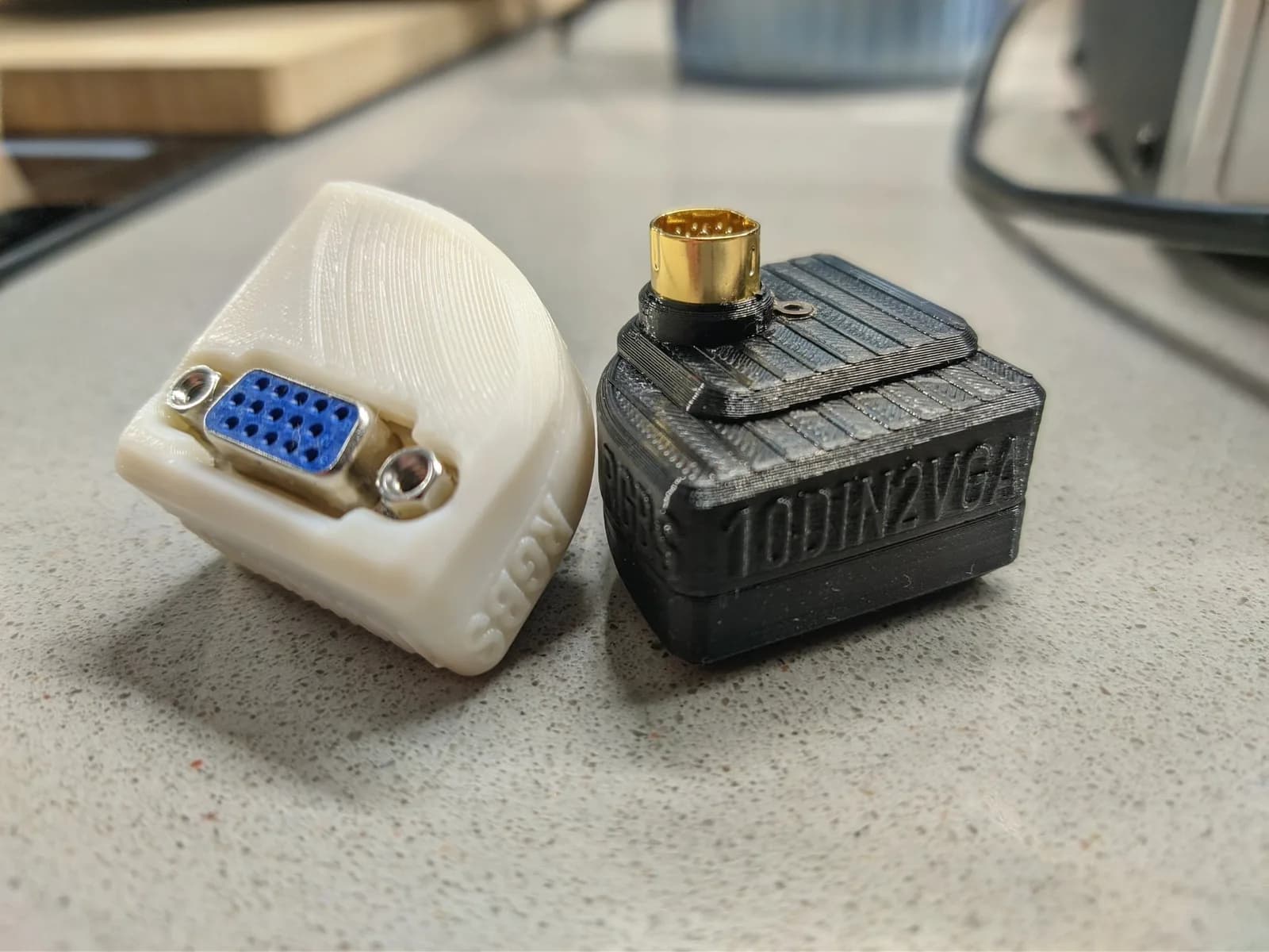 Two shelled 10DIN2VGA Saturn dongles: the white one (NTSC) showing its blue VGA connector and the black one (PAL, embossed '10DIN2VGA') showing the gold 10-pin DIN Saturn-side connector on top.
