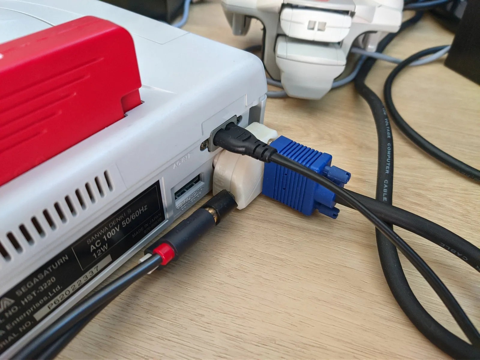 Close-up of the rear of a Japanese Sega Saturn (label 'SEGASATURN HST-3220') with a red Pseudo Saturn Kai cartridge in the top slot; the white/cream 10DIN2VGA dongle (the NTSC-configured build) on the A/V port feeds a blue VGA cable, and the AC mains cable is plugged into AC IN.