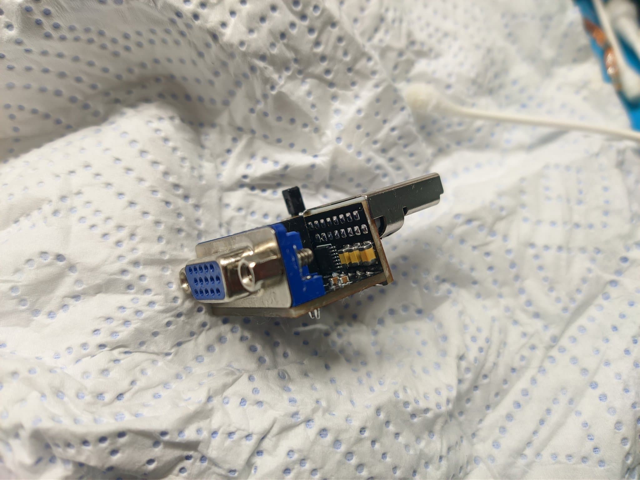 DC2VGA dongle on perforated mat showing blue PCB with IC chip, metal connector housing, and black plastic top cover.