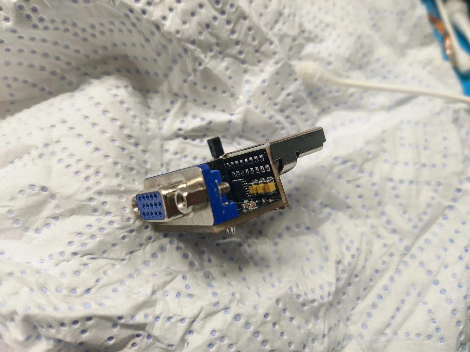 A DC2VGA (Dreamcast) dongle board on a paper towel, with its blue VGA connector, the metal Dreamcast A/V connector, an IC and brown MLCC capacitors, and the small black lever of the SD/VGA switch (which toggles 480p in VGA mode) on top.