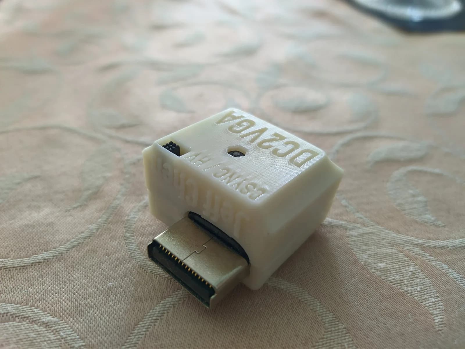 The DC2VGA dongle by Jeff Chen for the Dreamcast in a cream 3D-printed shell (embossed 'DC2VGA / Jeff Chen'), the mostly-silver metal Dreamcast connector protruding; a CSYNC/HV Sync toggle switch sits on the side of the case, on a cream patterned tablecloth.