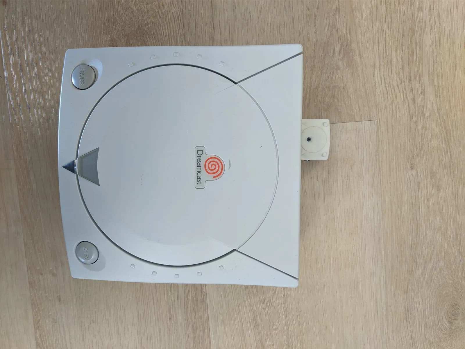 A Sega Dreamcast seen from above on a wooden surface, with the cream DC2VGA adapter connected to the A/V (video) port on the right side.
