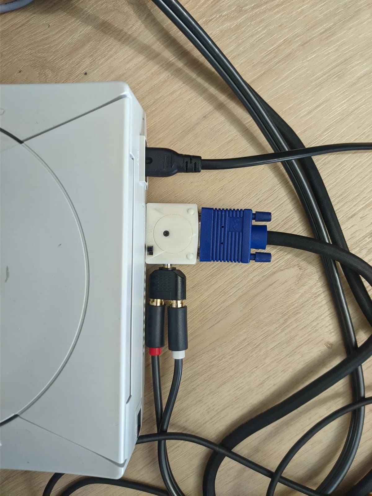 The white Sega Dreamcast with the DC2VGA dongle connected, showing the blue VGA connector, the power cable, and red/white RCA audio leads that feed through an adapter into the dongle's 3.5mm audio jack, on a wooden surface.
