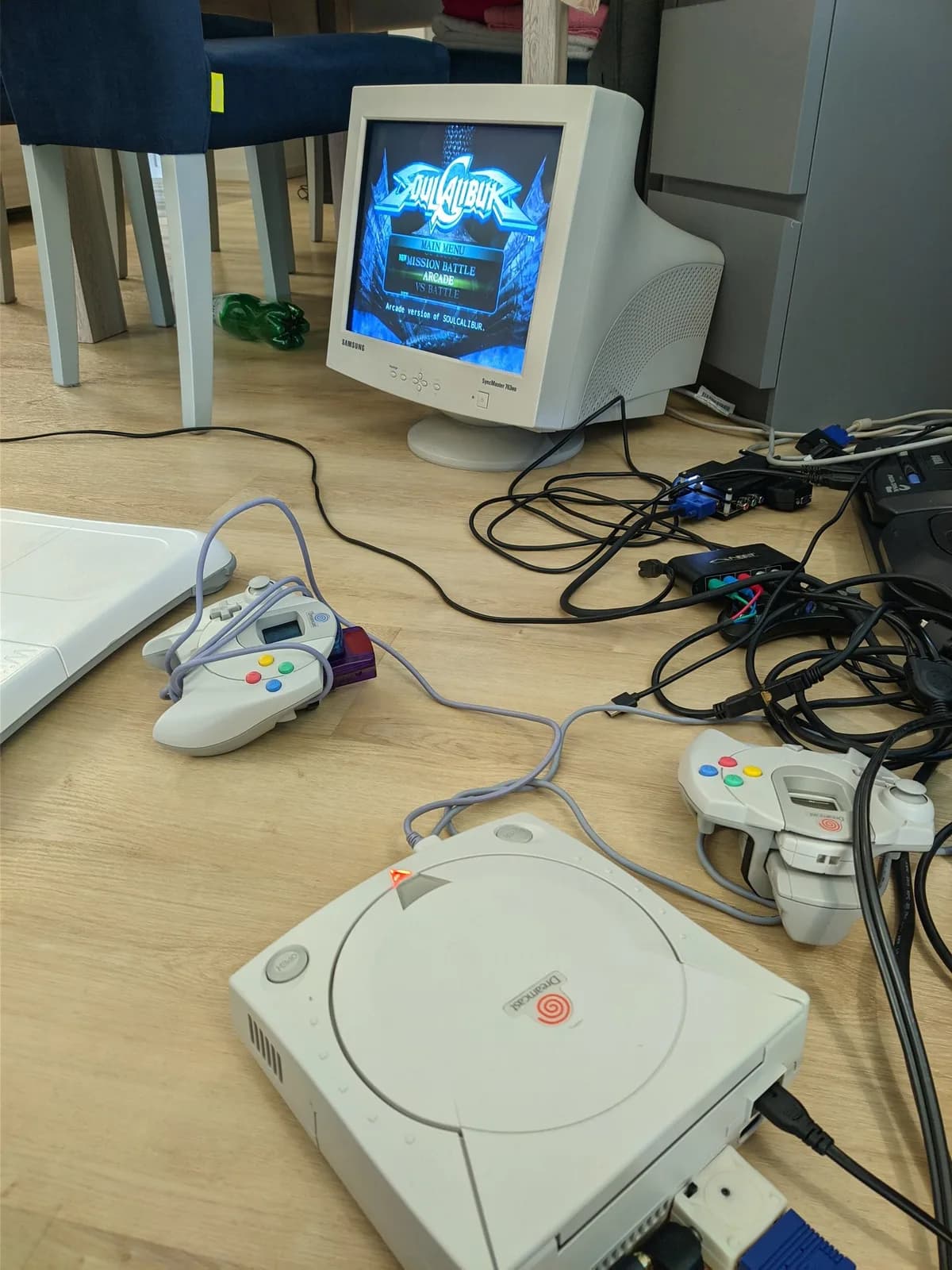 A Sega Dreamcast (powered on) with two controllers on a wooden floor, connected via the DC2VGA dongle to a Samsung SyncMaster 763MB CRT showing the Soul Calibur main menu (Mission Battle / Arcade / VS Battle).