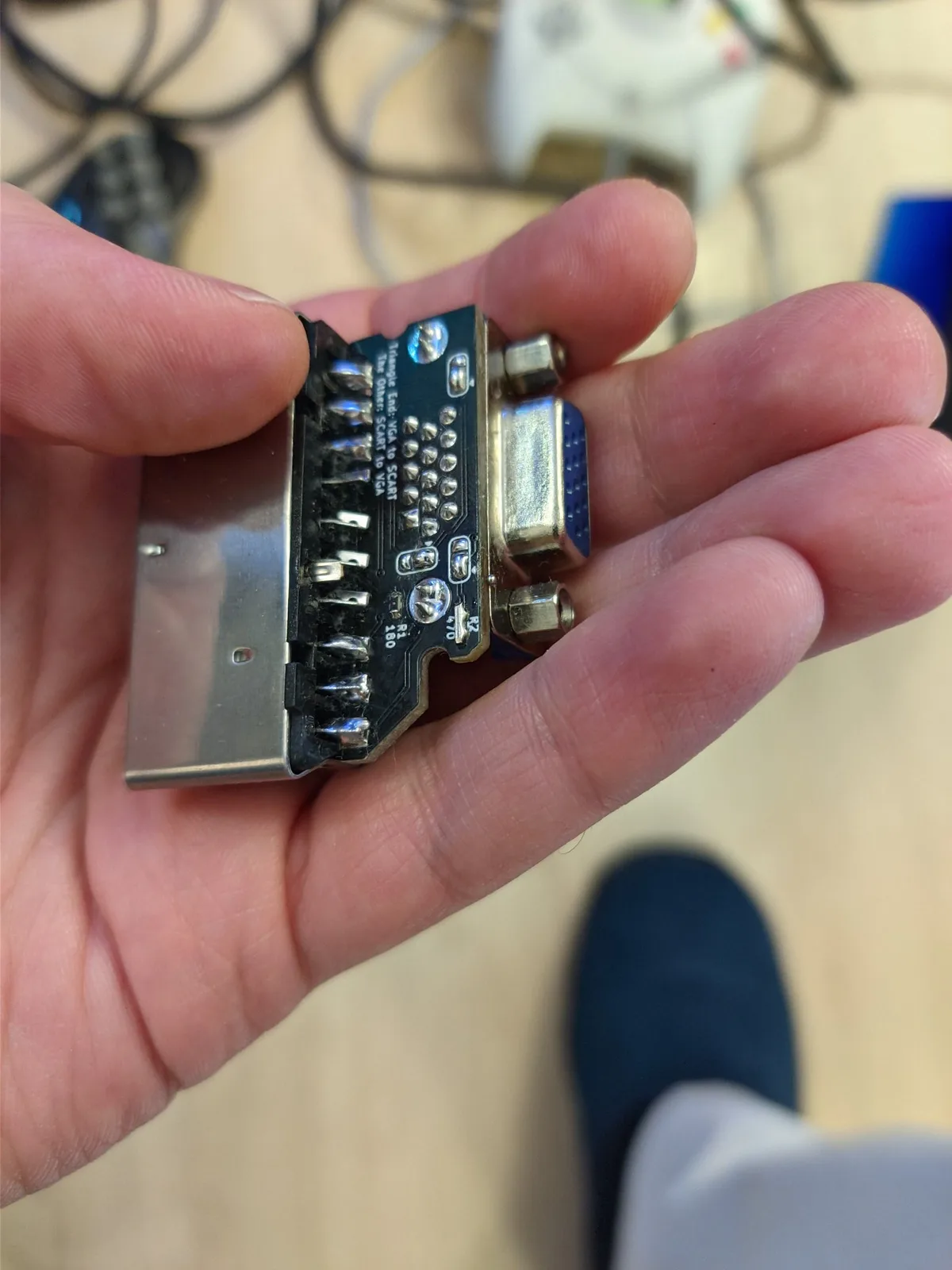 A hand holding the underside of the Jeff Chen VGAxSCART adapter board (metal connectors, black board, silkscreen 'VGA to SCART / SCART to VGA'), showing its configuration: the jumper is set to the triangle end and a resistor pad is bridged with solder and a metal link, setting it as VGA-to-SCART.