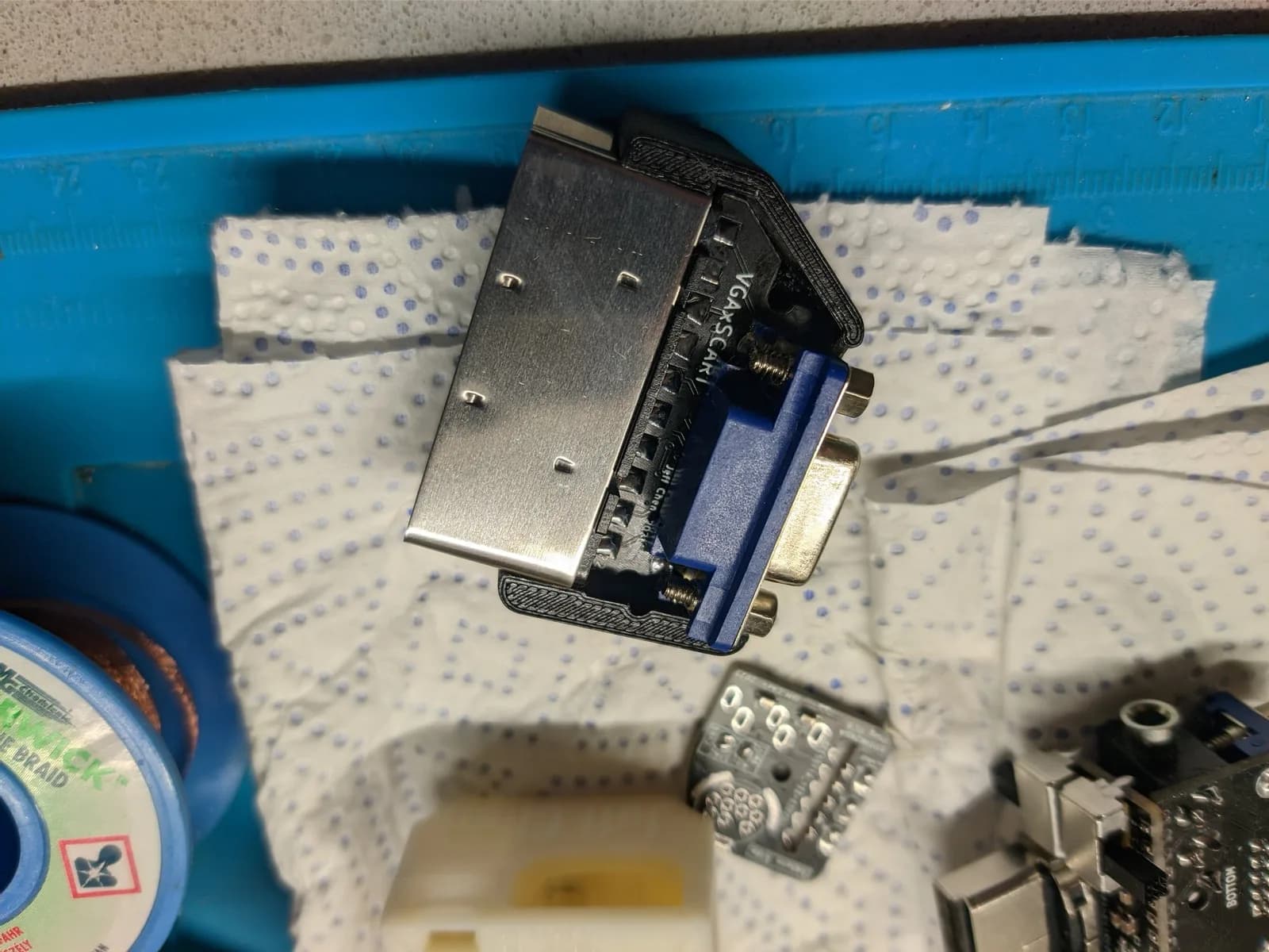 The Jeff Chen VGAxSCART adapter installed into one half of its black 3D-printed shell (the 'final form'), its blue VGA connector and metal SCART connector exposed, resting on a paper towel over a blue silicone work mat, with a spool of desoldering braid at left.