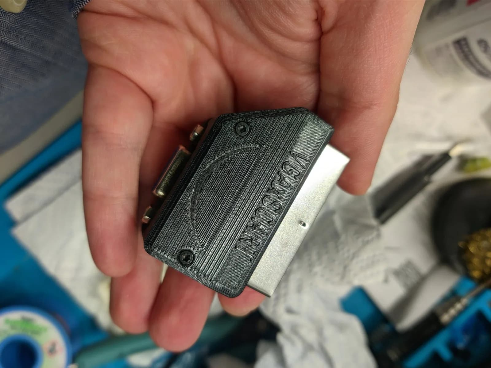 A hand holding the VGAxSCART adapter in its fully closed black 3D-printed shell, both halves screwed down, embossed 'VGAxSCART' with the Jeff Chen logo and the metal SCART connector protruding.
