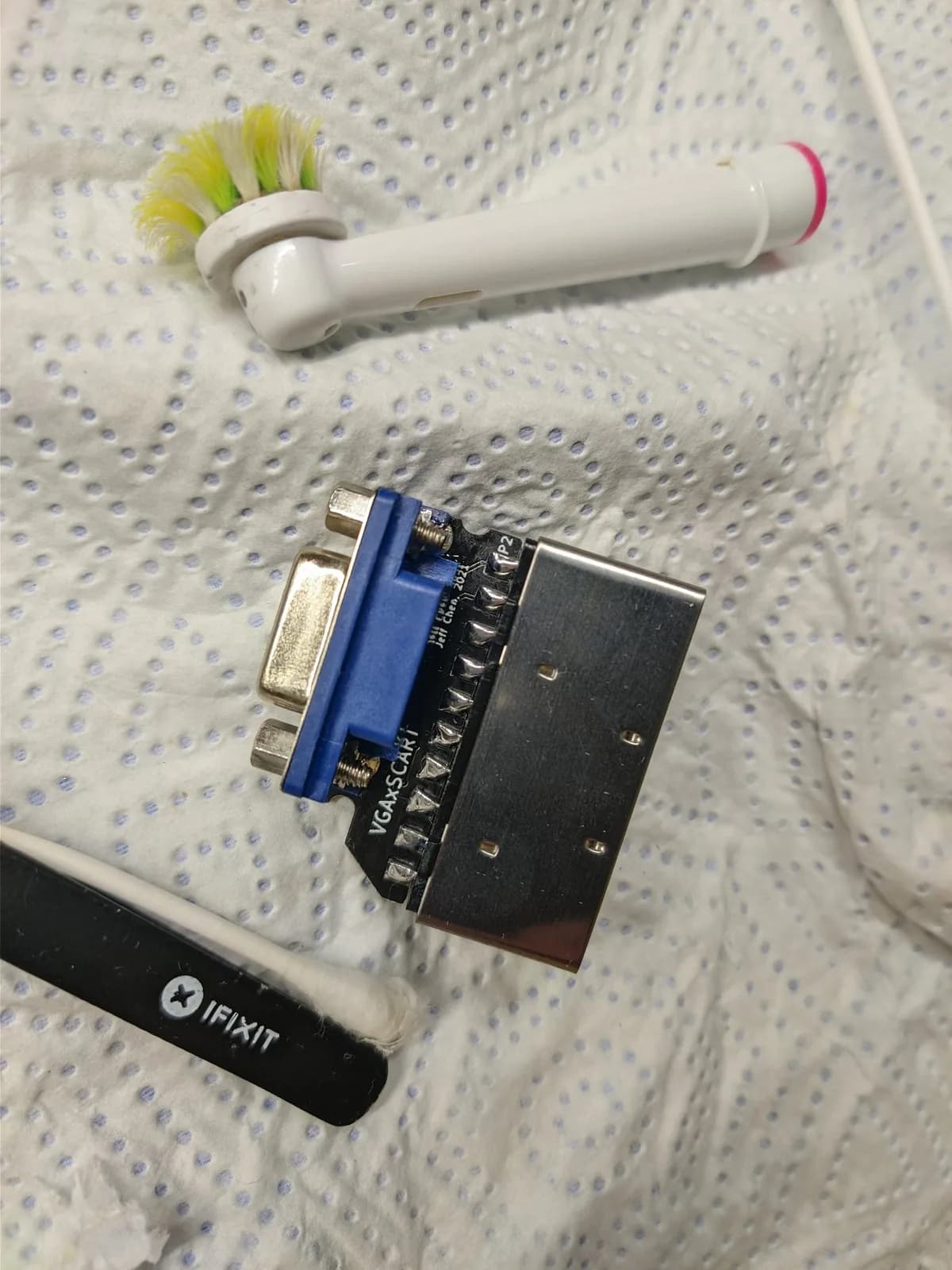 The top side of a Jeff Chen VGAxSCART adapter board (blue VGA connector, metal SCART connector, 'VGAxSCART / Jeff Chen' silkscreen) on a paper towel, with an electric toothbrush head used as a cleaning brush and a black iFixit tool and cotton swab beside it.