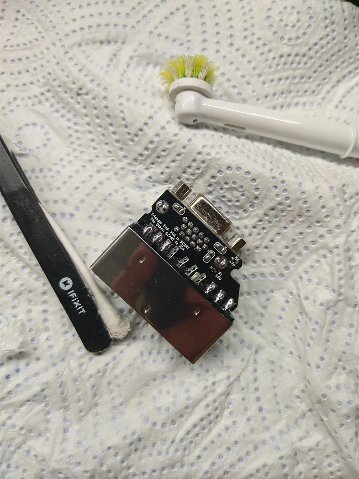 The underside of the Jeff Chen VGAxSCART adapter board on a paper towel, showing how it is configured (silkscreen 'Triangle End: VGA to SCART / The Other: SCART to VGA'); its blue VGA connector and metal SCART connector, with an electric toothbrush head and a black iFixit tool and cotton swab alongside.