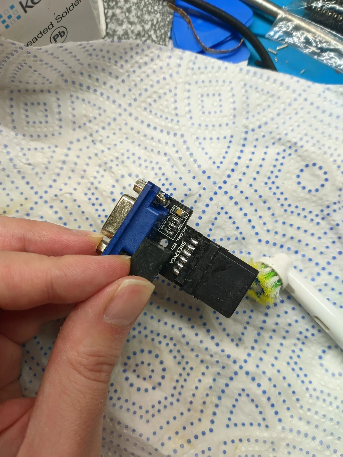 A hand holding the new-revision SNES2VGA dongle board, top side up ('SNES2VGA / Jeff Chen', blue VGA connector, black SNES multi-out plug), showing how it is configured, over a paper towel with a leaded-solder box and soldering iron behind.
