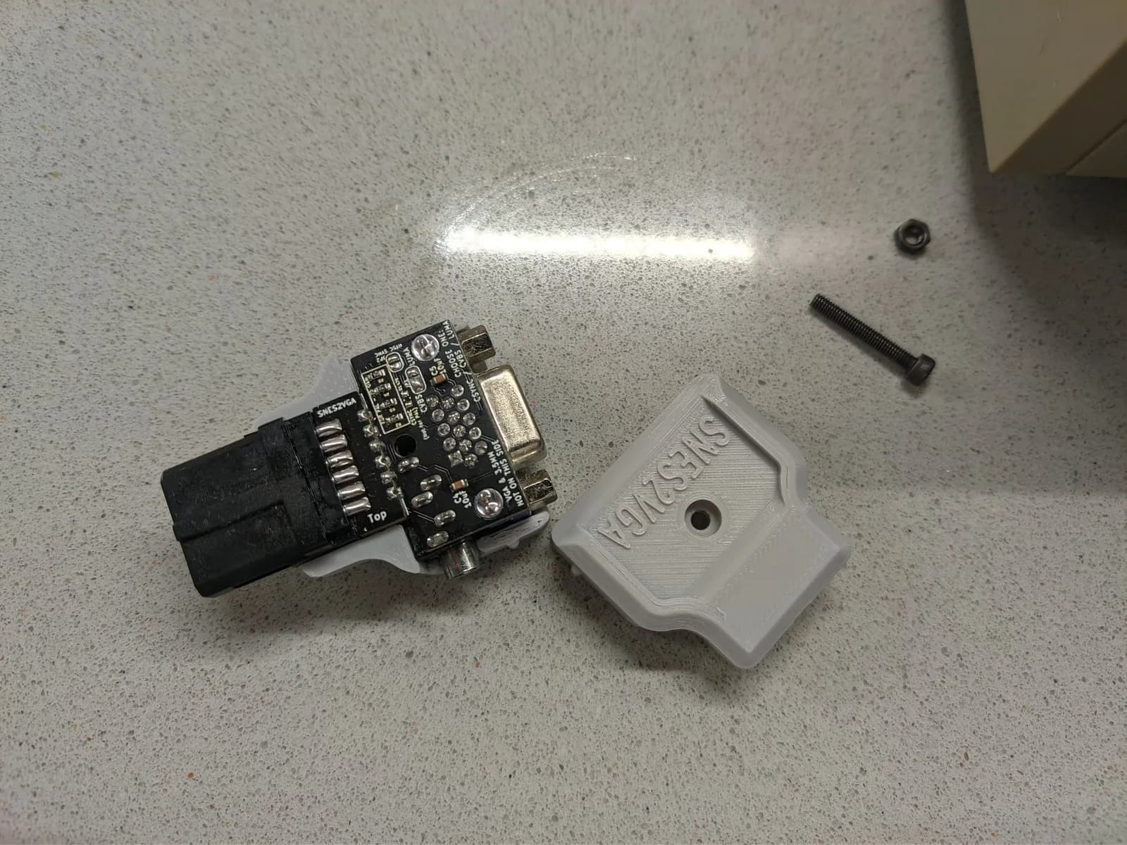 The bottom side of the new-revision SNES2VGA board on a speckled stone worktop, showing how it is configured (sync-select pads CVBS / CSYNC / Luma, silkscreen 'SNES2VGA'), beside its gray 3D-printed shell and a screw and nut; this is the revision Jeff Chen reworked to fix a PAL-console issue that André helped identify.