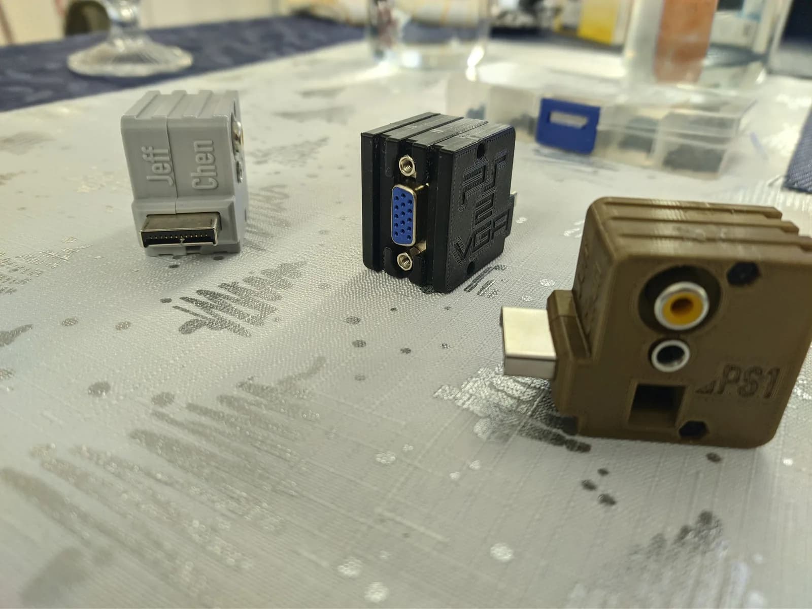 Three Jeff Chen VGA dongles on a mostly-white cloth patterned with silver Christmas-tree drawings: gray PS1-only (left), black PS2/PS3 with a blue VGA port (center), and a gold/tan universal (right).