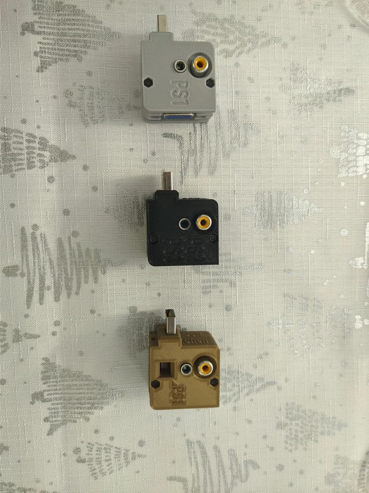 Top-view portrait shot of three Jeff Chen PlayStation VGA dongles laid out on a mostly-white cloth with silver Christmas-tree drawings: gray PS1-only, black PS2/PS3, and gold universal; each has a PlayStation multi-out plug plus a composite RCA jack and a 3.5mm jack, and the gray one shows its blue VGA output.