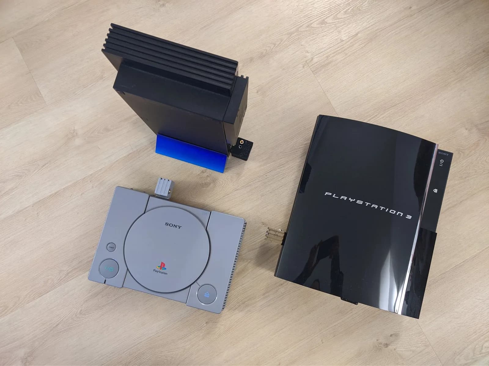 Three PlayStation consoles on a wooden floor, each wearing its matching Jeff Chen dongle: a gray PS1 (gray dongle), a black PS2 'fat' standing on a blue stand (black dongle), and a black PS3 'fat' labelled PLAYSTATION 3 (gold dongle).