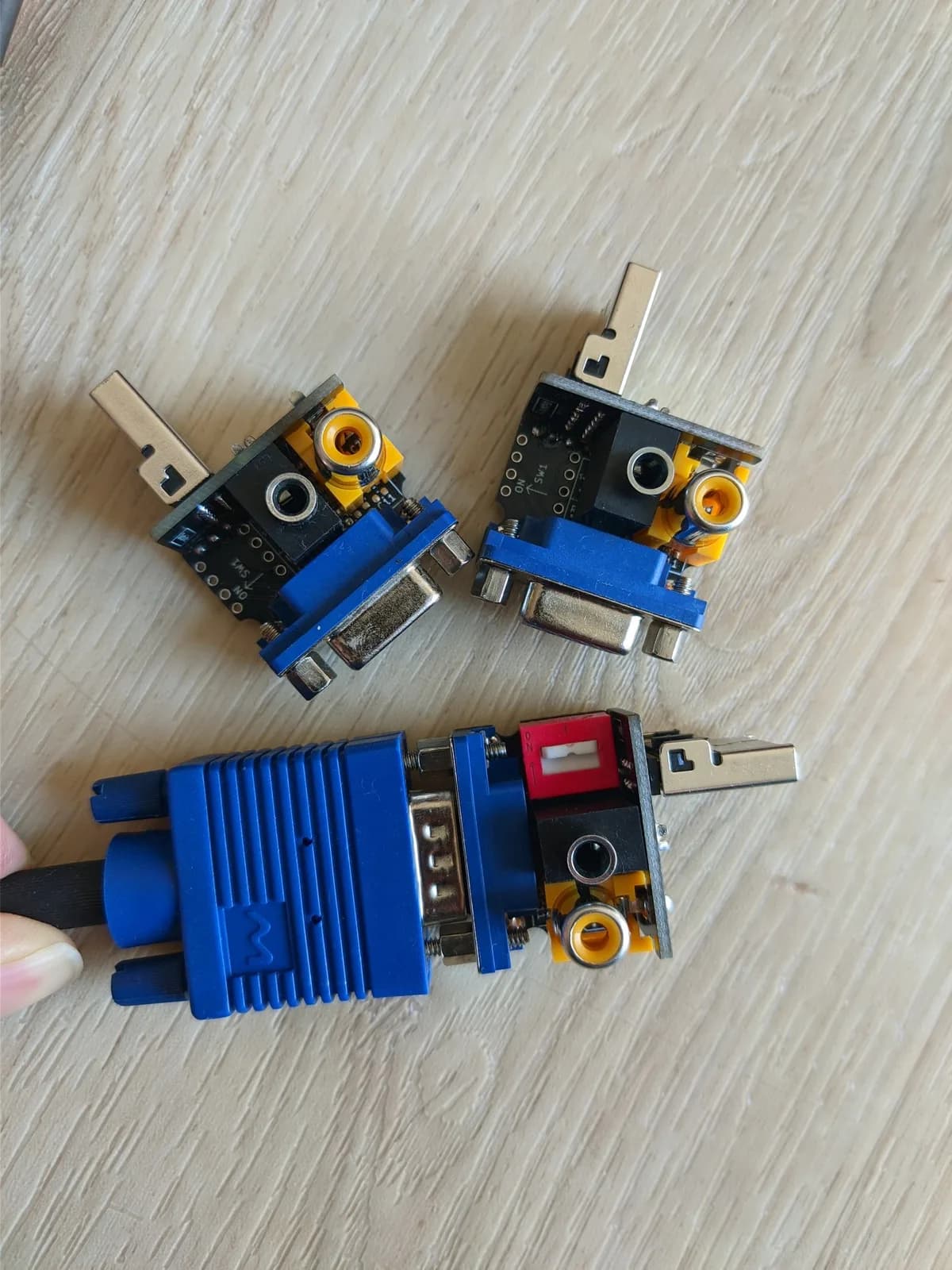 Three PS2VGA dongles on a wooden surface, each with a blue VGA connector, a yellow RCA composite jack (for lightgun support), a 3.5mm jack, and the metal PlayStation multi-out plug; one has a red DIP switch (SW1) and the front one has a blue VGA cable plugged into it.