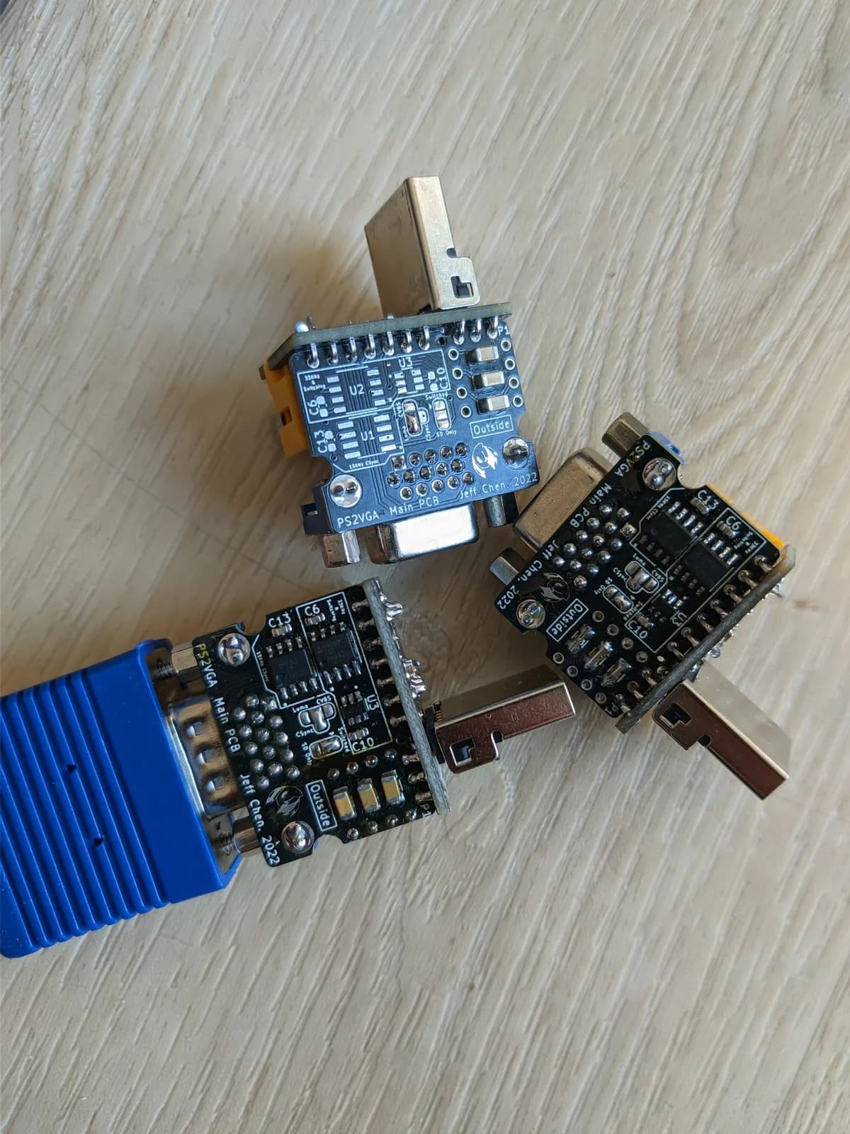 The undersides of three PS2VGA dongle boards on a wooden surface ('PS2VGA Main PCB / Jeff Chen 2022' silkscreen), showing the component placements and jumper settings for the different build configurations; the PS1-only version has noticeably fewer ICs populated. Metal PlayStation multi-out plugs and one blue VGA connector are visible.