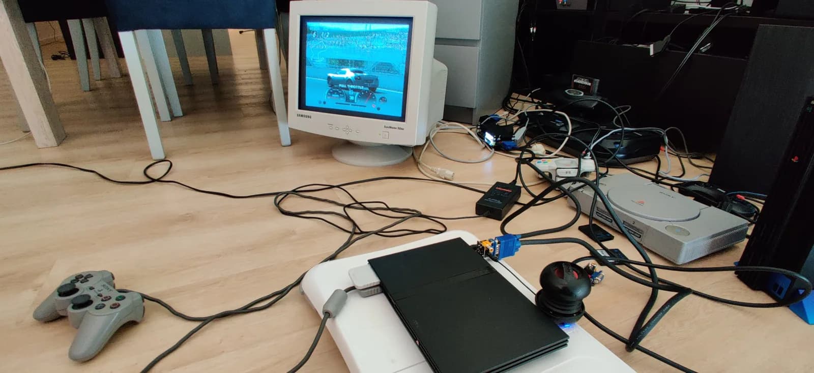 A black PlayStation 2 slim resting on a white Wii Balance Board, connected through a Jeff Chen VGA dongle to a Samsung SyncMaster 763MB CRT showing Gran Turismo 4; a gray Sony PlayStation controller lies in the foreground on the wooden floor.