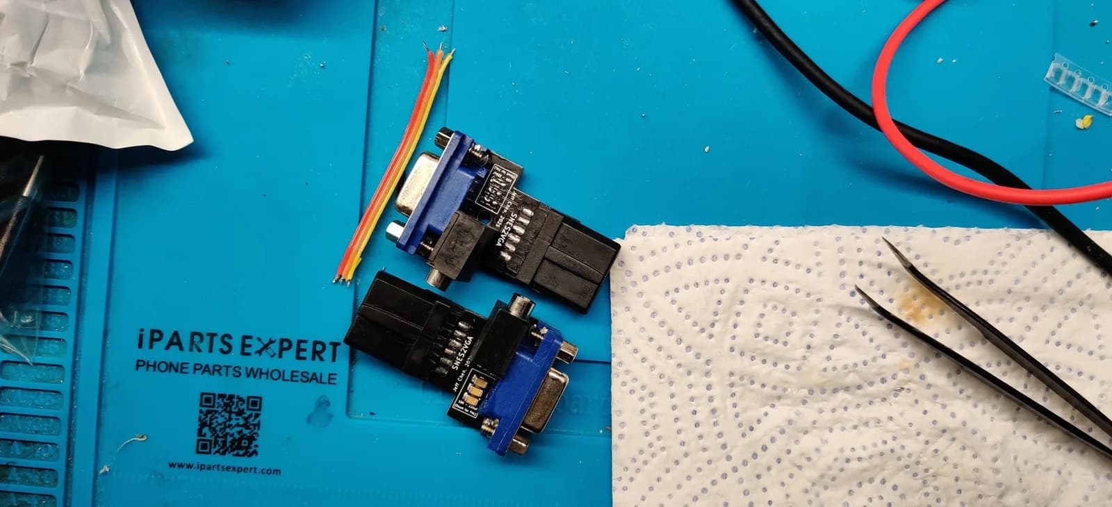 Two assembled SNES2VGA dongles (blue VGA connectors, black SNES multi-out plugs, 'SNES2VGA / Jeff Chen' silkscreen) on a blue iParts Expert silicone mat, one configured for PAL consoles and the other for NTSC; red and black multimeter leads, loose coloured hookup wires, and tweezers lie on a paper towel alongside.
