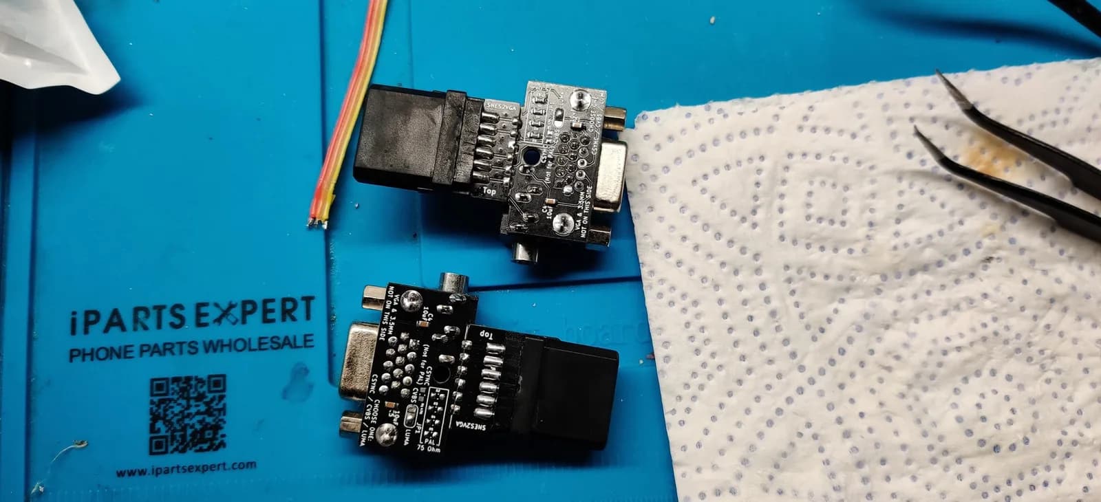 The undersides of the same two SNES2VGA dongles from the previous photo, on a blue iParts Expert mat, showing the sync-select solder pads (CSYNC / CVBS / LUMA) and 'SNES2VGA / Jeff Chen' silkscreen; loose coloured hookup wires and tweezers nearby.