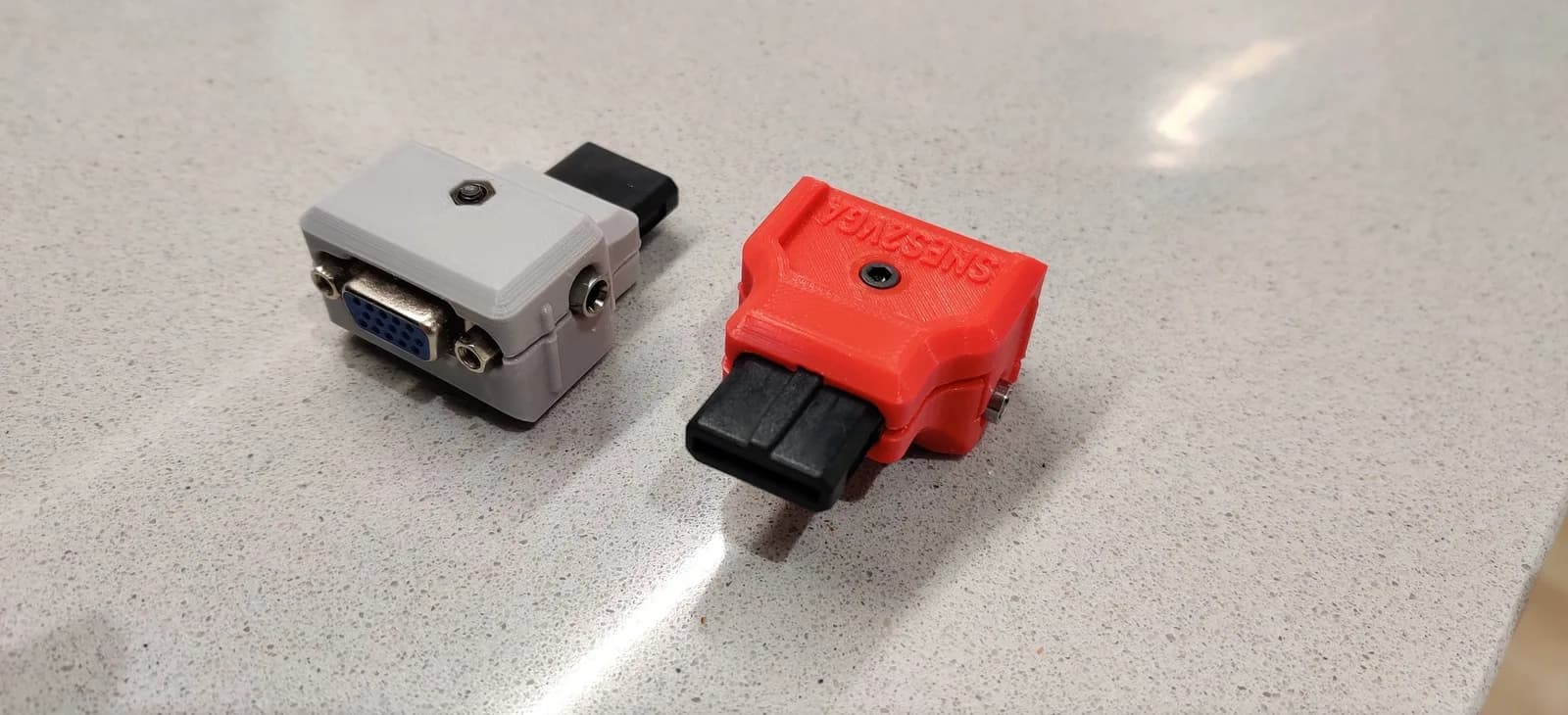 Two SNES2VGA dongles on a speckled stone worktop: the grey one (configured for PAL consoles) showing its blue VGA connector, and the red one (configured for NTSC, silkscreen 'SNES2VGA') showing the black SNES multi-out plug; both have a small 3.5mm jack.