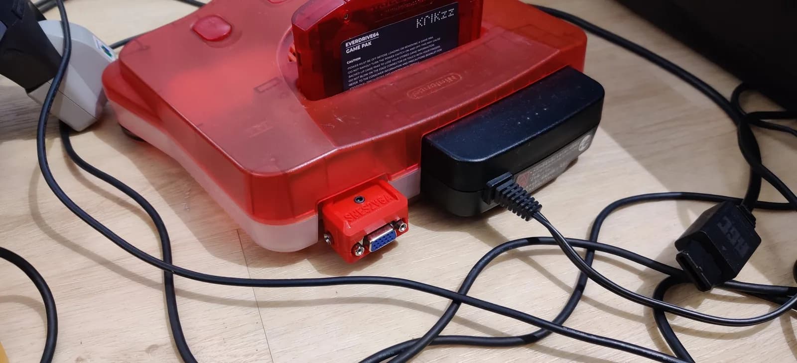 A Japanese (NTSC) Nintendo 64 with a clear-red top and frost-white base on a wooden surface, a red-shelled SNES2VGA dongle (the NTSC-configured build) plugged into its multi-out and an EverDrive 64 flashcart (krikzz) in the cartridge slot; a black power supply and cables sit alongside.