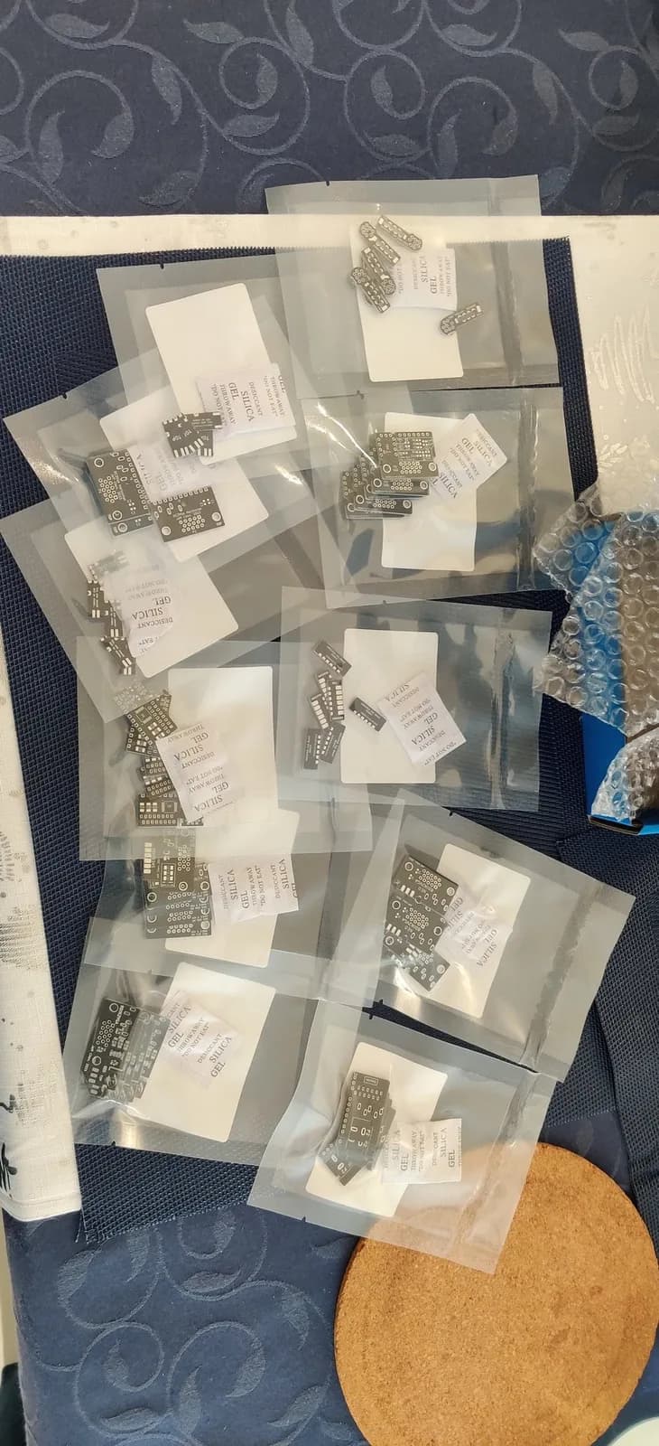 A pile of bare Jeff Chen dongle PCBs (well over a dozen), each sealed in a clear anti-static bag with a silica-gel desiccant packet, on a dark blue patterned tablecloth with bubble wrap nearby; the boards are unpopulated, no SMD components or pin headers fitted yet, five PCBs to a bag for each dongle type.