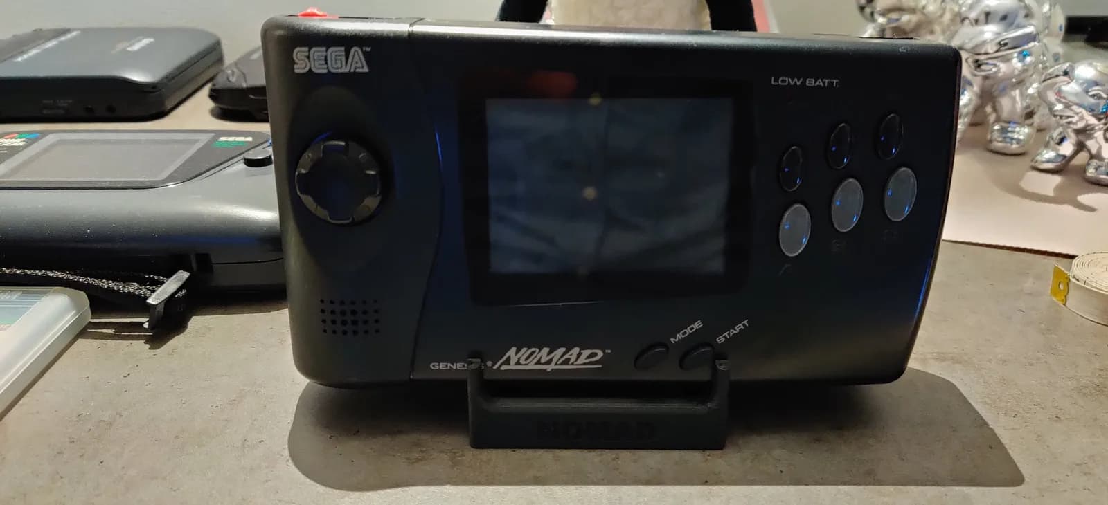 Sega Nomad handheld console on display stand with restored screen, surrounded by other gaming devices and collectibles on workbench.