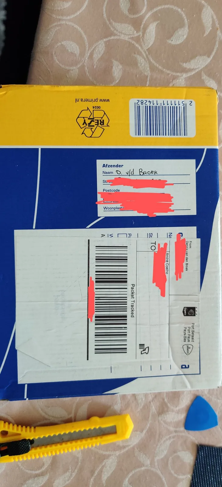 The cardboard shipping box the gifted PSOne arrived in, a blue and yellow Dutch parcel box on a patterned cream tablecloth, its sender and recipient address labels scribbled out in red marker, with a yellow snap-blade utility knife and a blue plastic pry tool lying nearby.