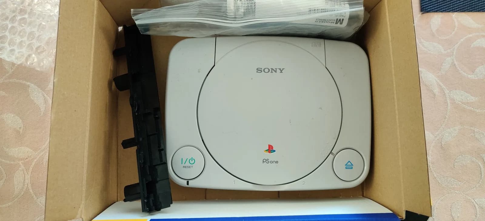 The gift shipment in a brown cardboard box: a black Panasonic Q optical-drive rail and a clear Mouser Electronics bag holding the shattered second Q rail, with the gifted white Sony PSOne (SCPH-101) in the box alongside them.