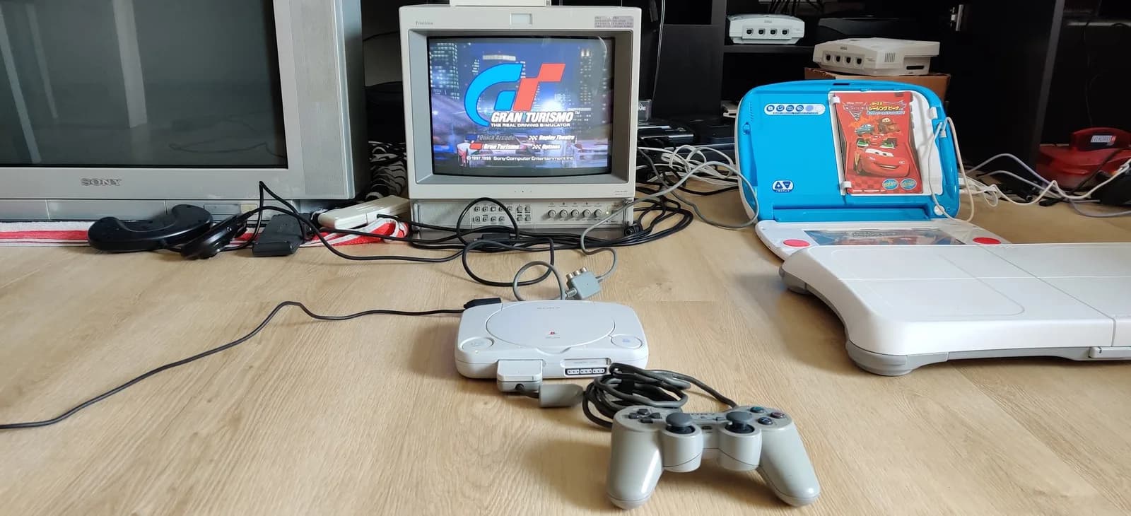 White Sony PSOne (SCPH-101) with a grey DualShock controller on a wood floor, the original PlayStation Gran Turismo title menu on a Sony PVM-14L2, more retro consoles and a larger Sony KV-29FX30E Trinitron around the setup.