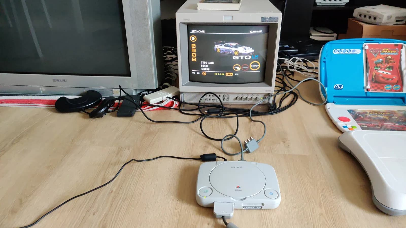 White Sony PSOne (SCPH-101) with its AV cable on a wood floor, connected to a Sony PVM-14L2 showing the Gran Turismo garage screen with a silver Mitsubishi GTO LM race car.