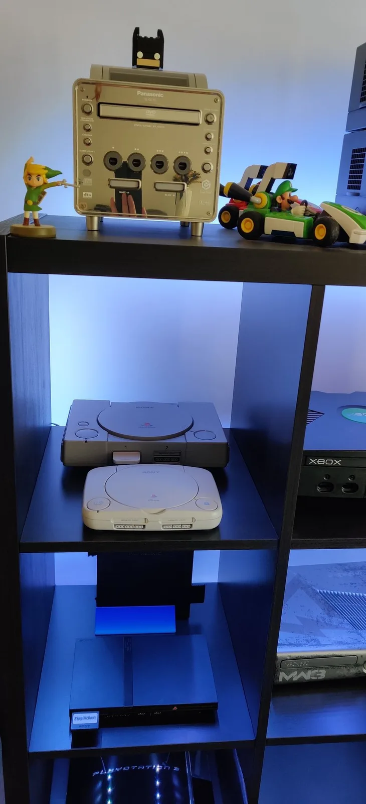 The recapped white PSOne (SCPH-101) back in its place on a blue-LED-lit black cube shelf of retro consoles, an original grey PlayStation immediately behind it on the same cube, a black original Xbox to the right and a silver, mirror-fronted Panasonic Q up top.
