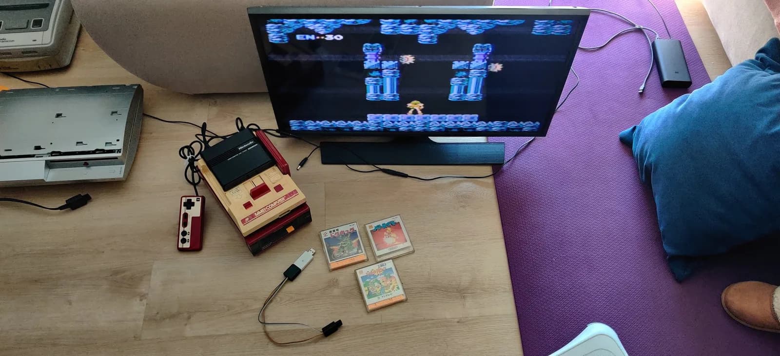 Famicom Disk System with homemade FDSStick transfer cable connected, three game cartridges displayed, retro gaming setup with monitor showing gameplay.