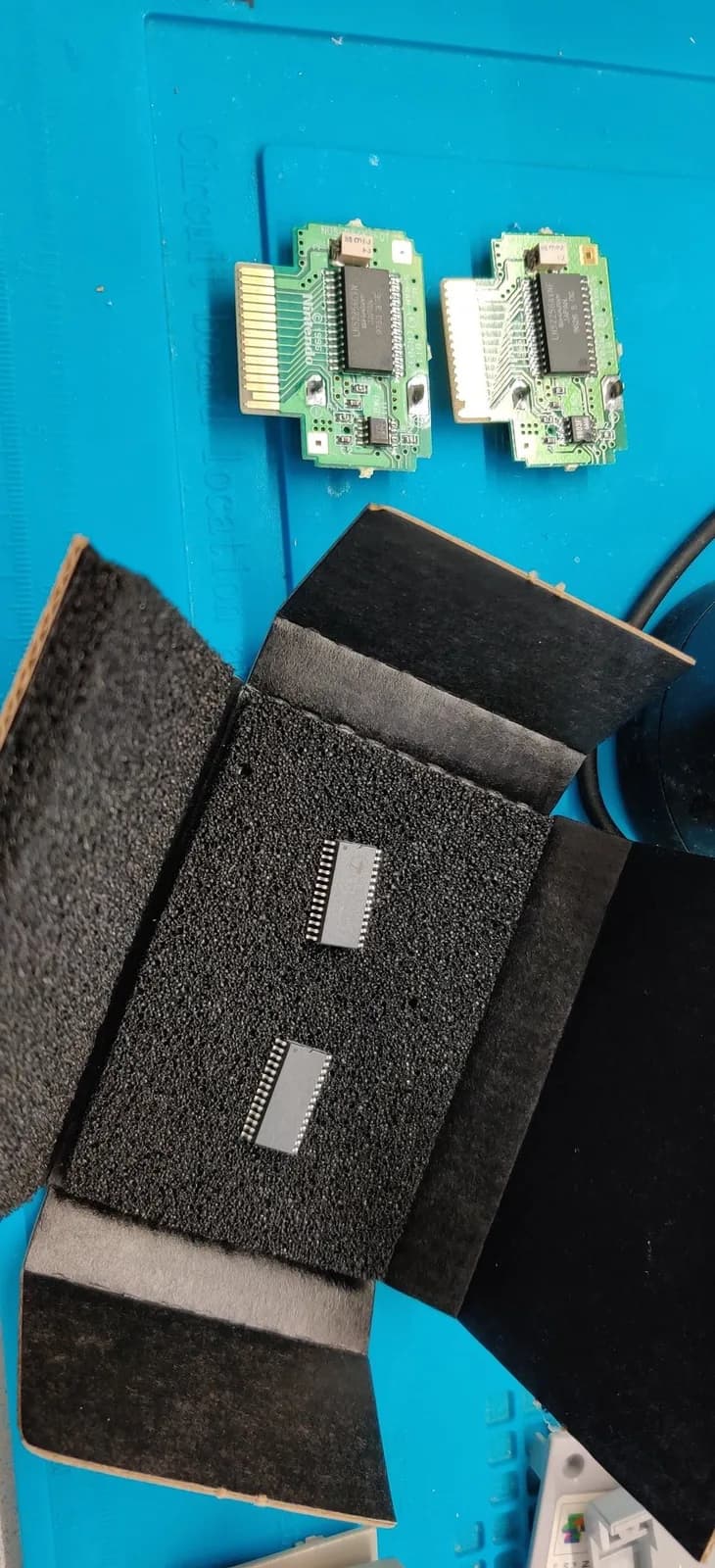 Two N64 Controller Pak PCBs with F-RAM chips installed, displayed above foam storage case containing additional memory modules.