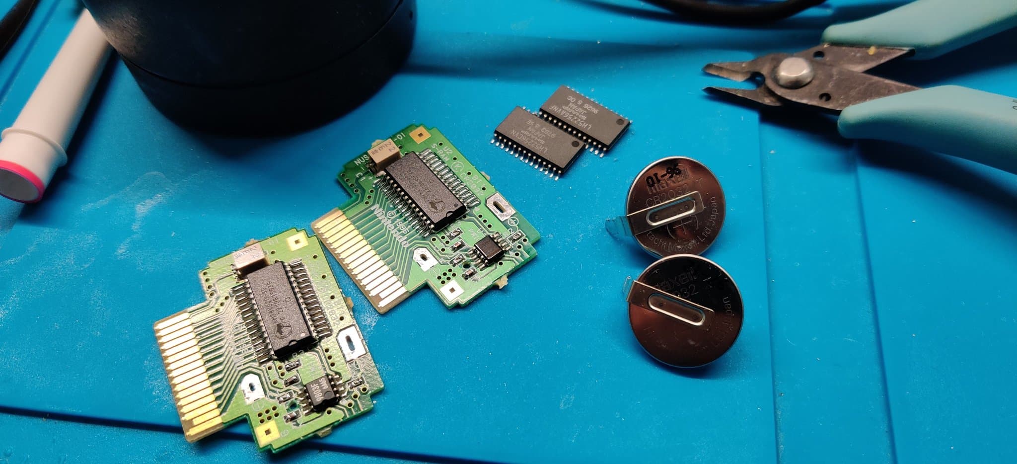 Two N64 Controller Pak PCBs with F-RAM chip upgrades, loose ICs, pennies for scale, and repair tools on blue mat.