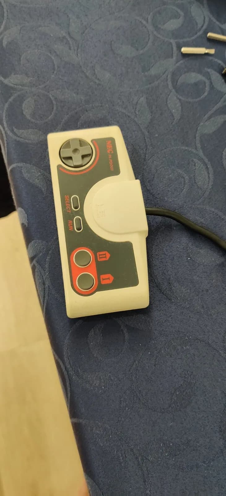 Cream and dark gray retrobrited game controller on patterned fabric, showing improved plastic coloring from yellowing restoration treatment.