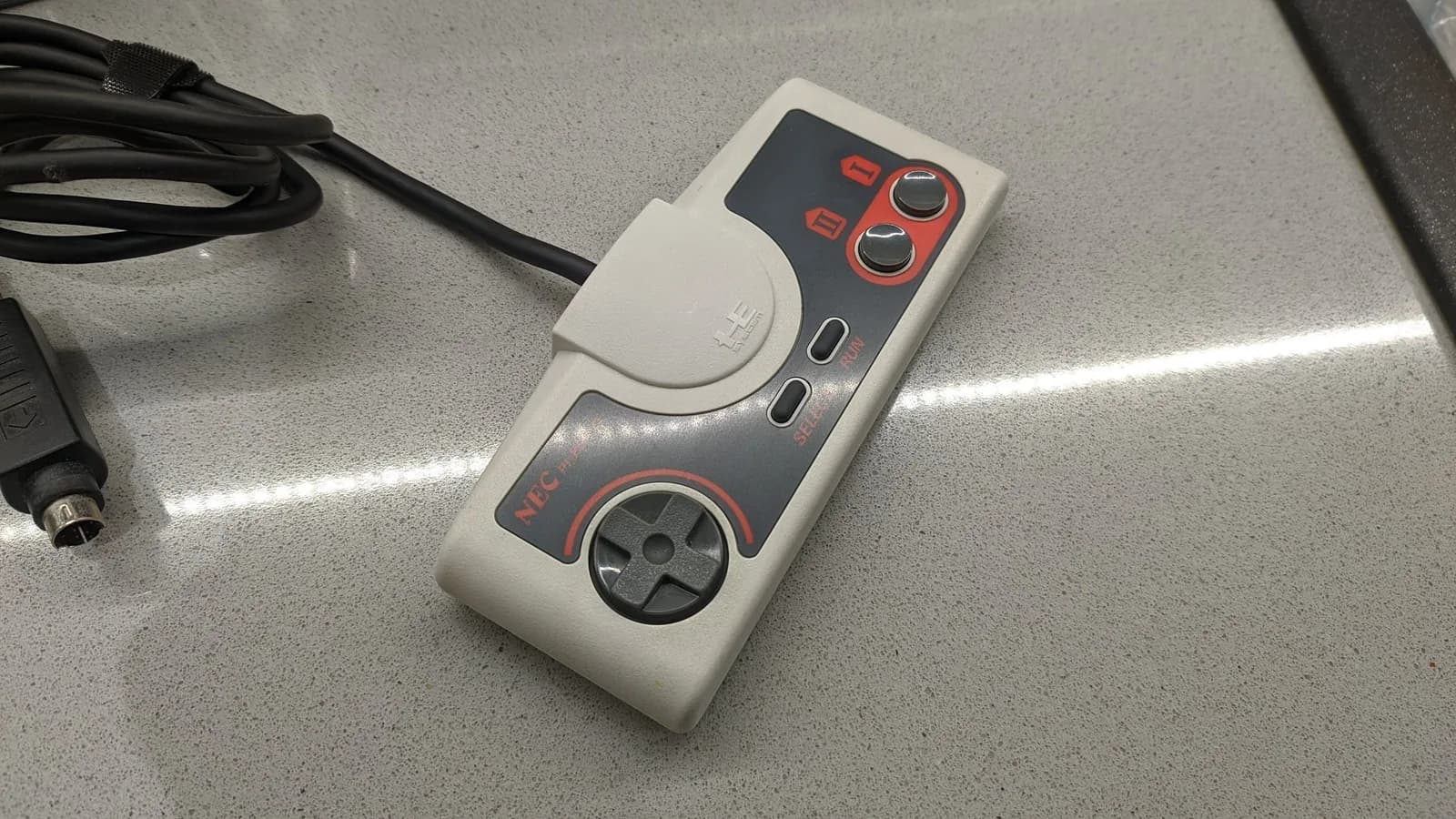 Retrobrited PC Engine controller with red and black accents on metallic surface, alongside coiled cable and power connector.