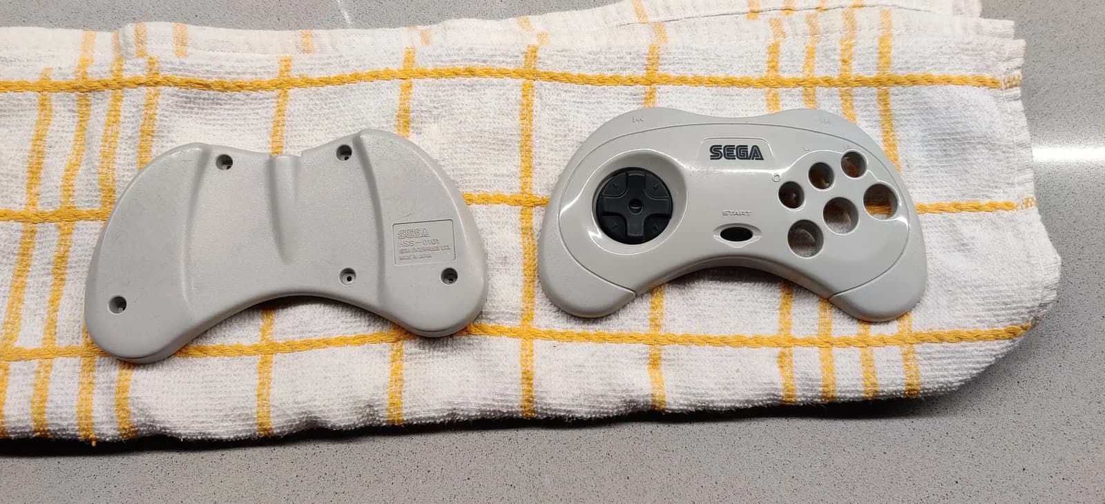 Two white Sega controllers on yellow-striped towel, showing back and front views after retrobriting treatment to remove yellowing.