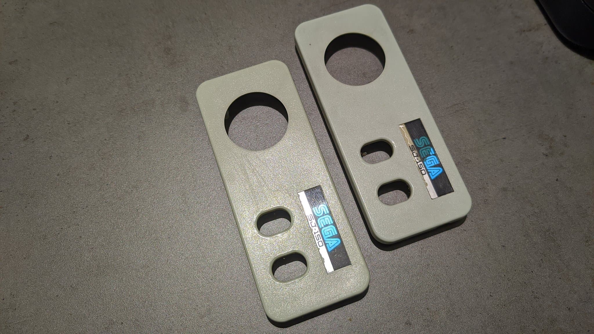Two cream-colored Sega SG-1000 II controllers after retrobrighting, showing brightened plastic with circular button holes and manufacturer labels visible.