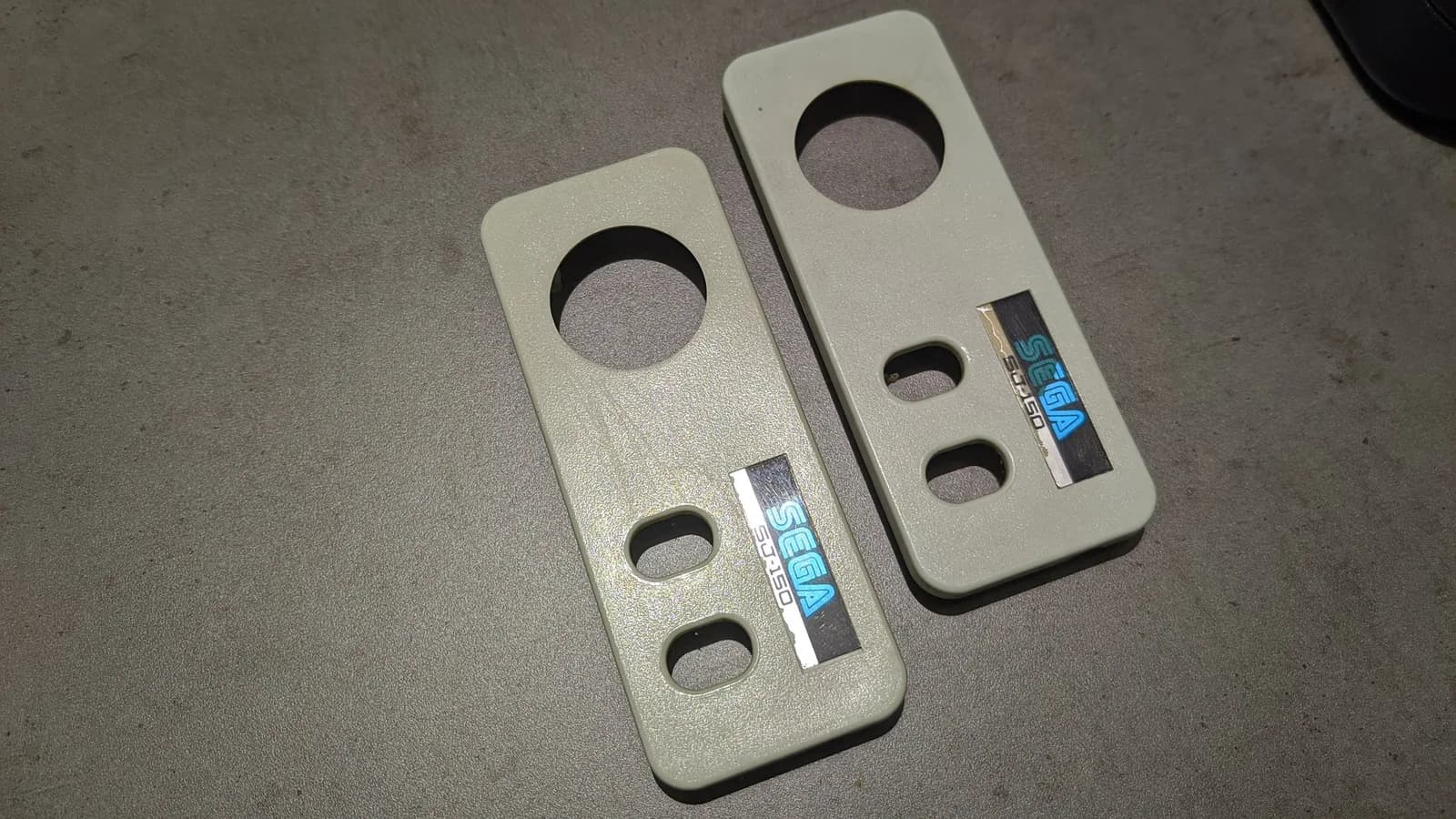Two Sega SG-1000 II controller faceplates on a dark surface, slightly yellowed (cream-coloured) before retrobriting, each with a D-pad cutout, two round button holes and a blue 'SEGA SG-1000' sticker.