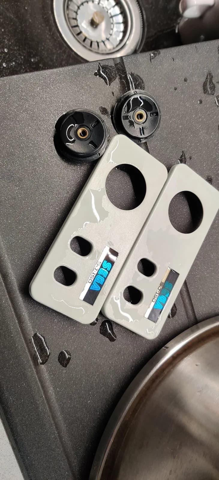 Two Sega SG-1000 II controller faceplates, wet after being washed at the end of a vapour-method retrobrite, resting in a sink beside the two black D-pads.