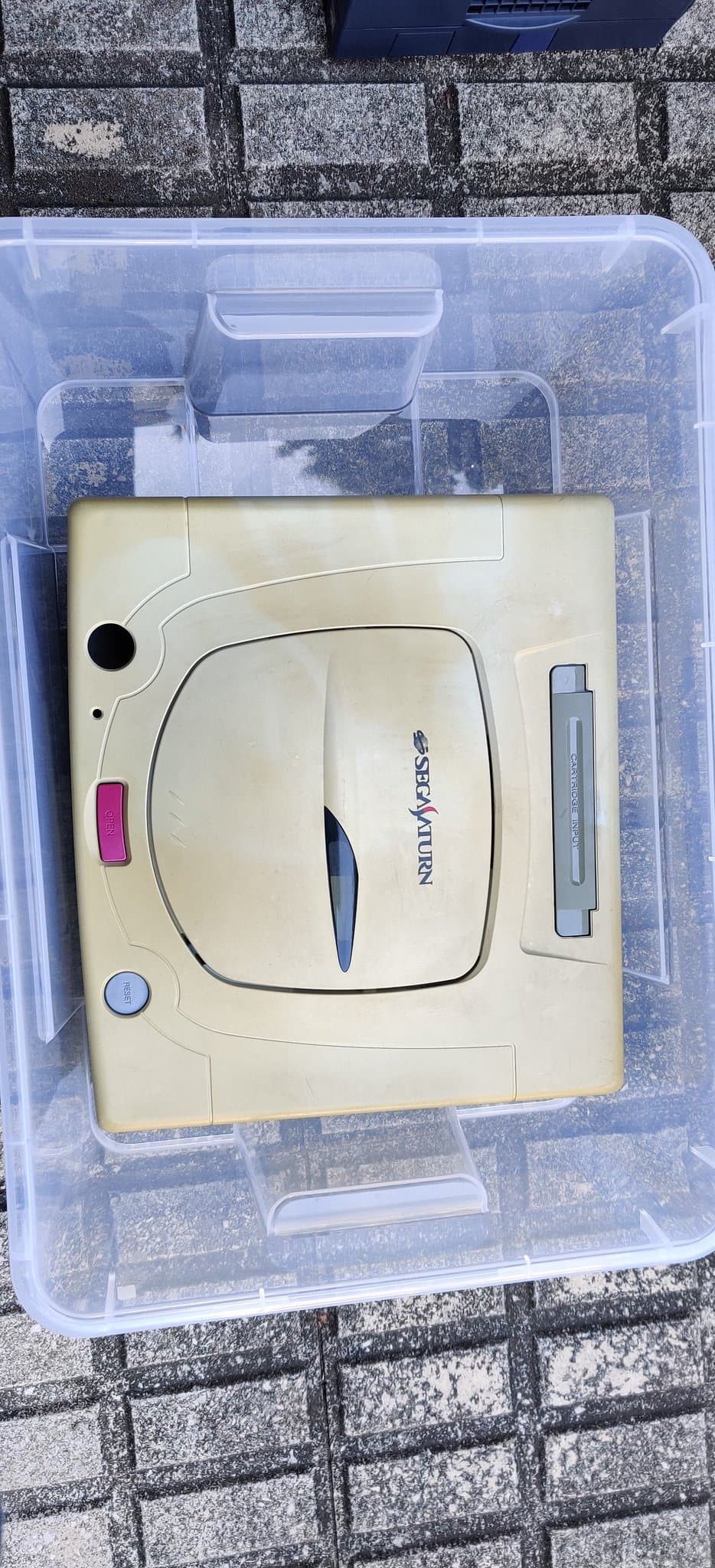 Cream-colored Sega Saturn model 2 in clear plastic storage bin showing retrobrighting progress with hydrogen peroxide treatment.