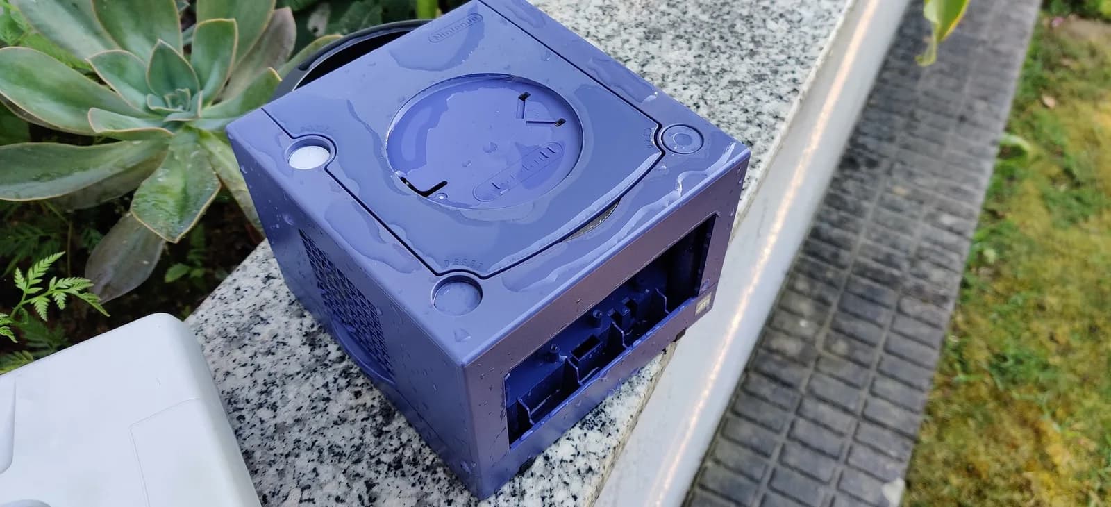 The indigo GameCube shell on a granite ledge outdoors after the retrobrite, rinsed with tap water and looking indigo again rather than the near-black it had darkened to (the controller-port panel is not mounted in this shot).