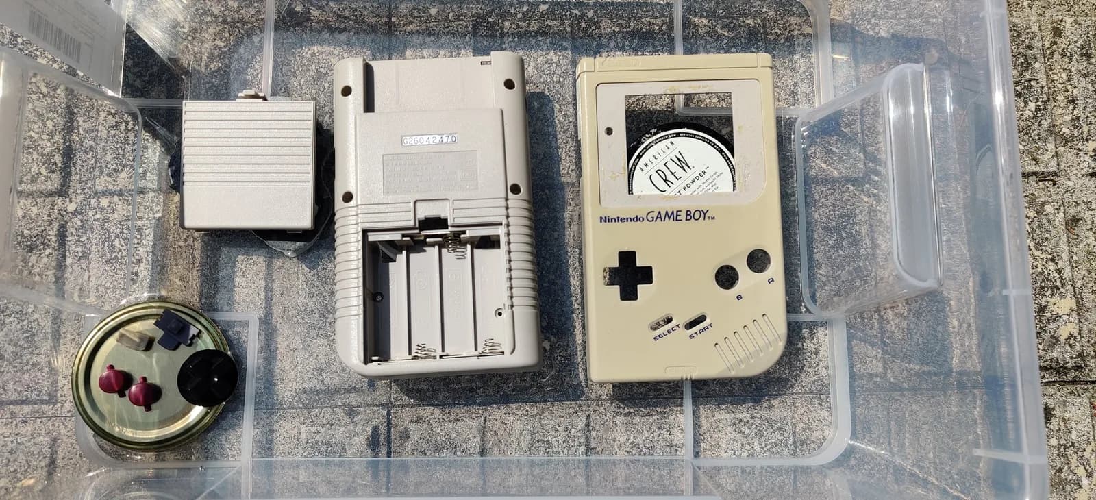 Original Nintendo Game Boy DMG, a companion piece retrobrited alongside the Mark III, disassembled in a clear container ready for the vapour-method retrobrite (a pre-process shot): the GAME BOY front shell with d-pad and buttons, the back shell with battery compartment, and small parts in a jar lid.