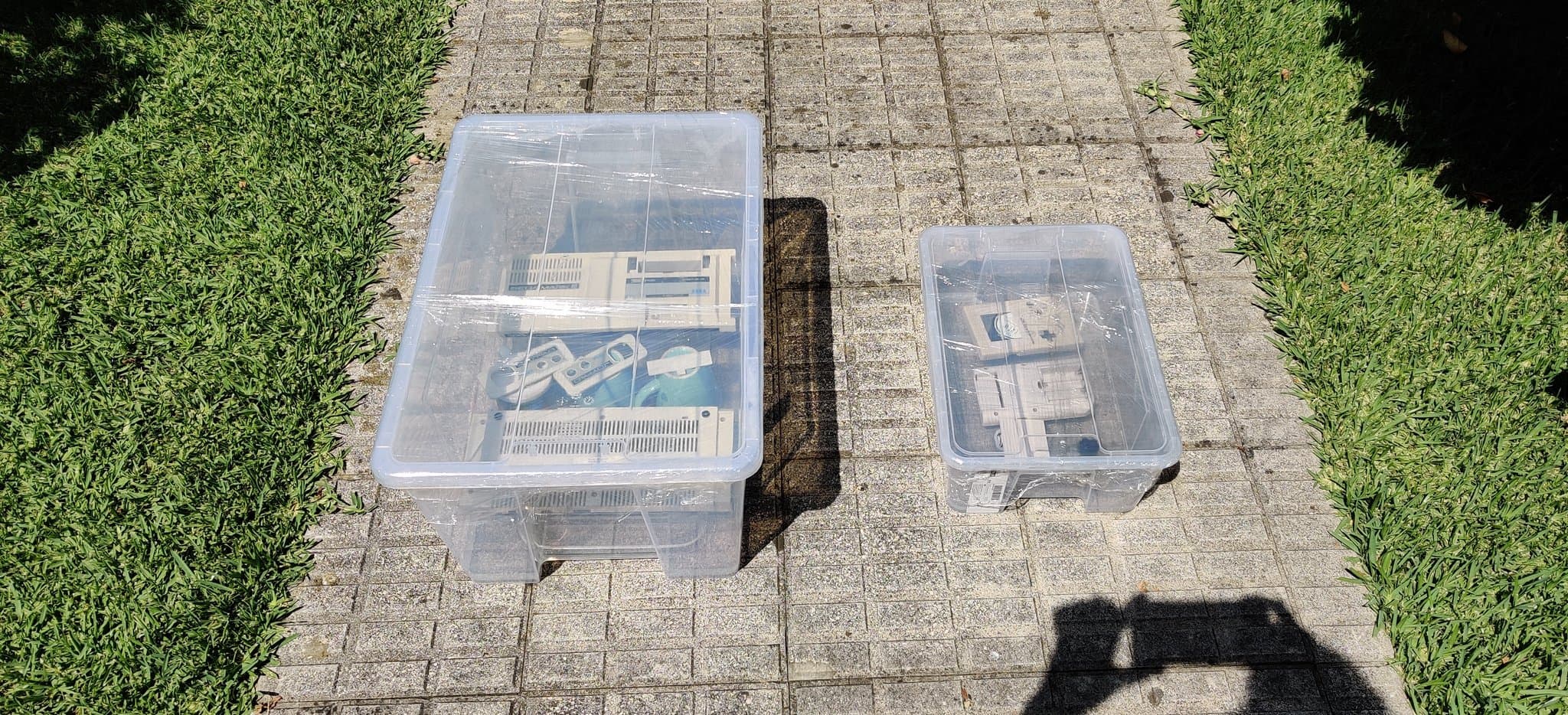 Two clear plastic storage containers on brick pavement holding Sega Mark III console parts during retrobrighting refurbishment process.