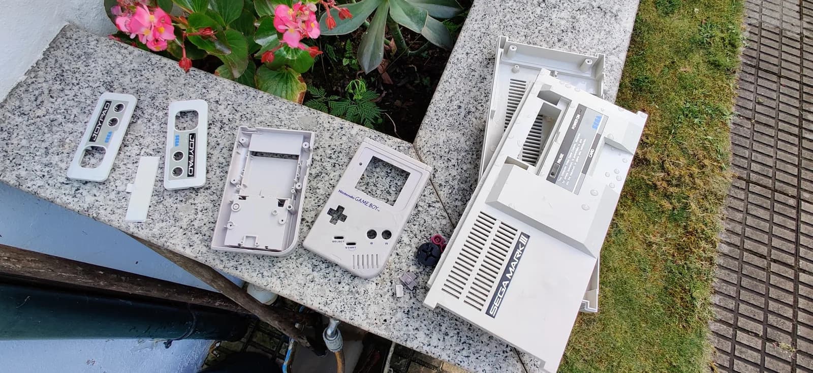 Retrobrited shells washed and laid out on a marble ledge with flowers behind, all looking much better than before: the full cream Sega Mark III shell (both halves, louvered vents) and its Joypad controller shells, alongside the Nintendo Game Boy DMG shell pieces.
