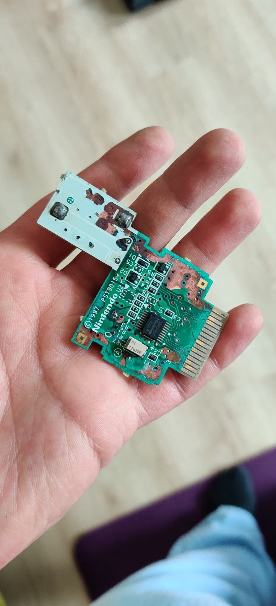 Hand holding the heavily corroded N64 Rumble Pak green circuit board before repair, component side up. The board carries its surface-mount parts including the black motor-driver chip and the gold edge connector, with exposed copper traces and corroded vias from the battery leak, and an oddly shaped white section that is part of the board itself.