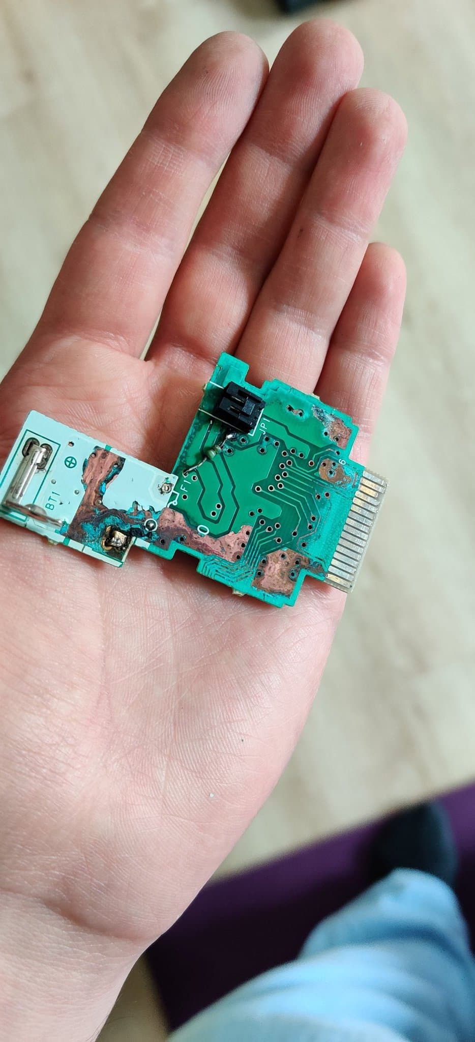 N64 Rumble Pak green circuit board held in a palm before repair, non-component side up. The battery contact area (marked BT1) on the white part of the board shows teal and copper battery-leak corrosion, the motor connector sits beside it, and a resistor has been relocated so the pak can draw power from the controller instead of the battery terminals. The gold edge connector is at the right.